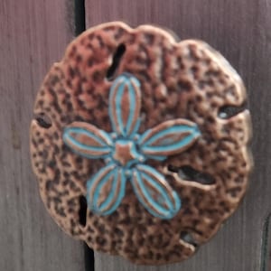 May include: A bronze-colored sand dollar-shaped decorative element with a turquoise and bronze flower design in the center. The sand dollar has a textured surface and several small holes. The design is likely for home decor.