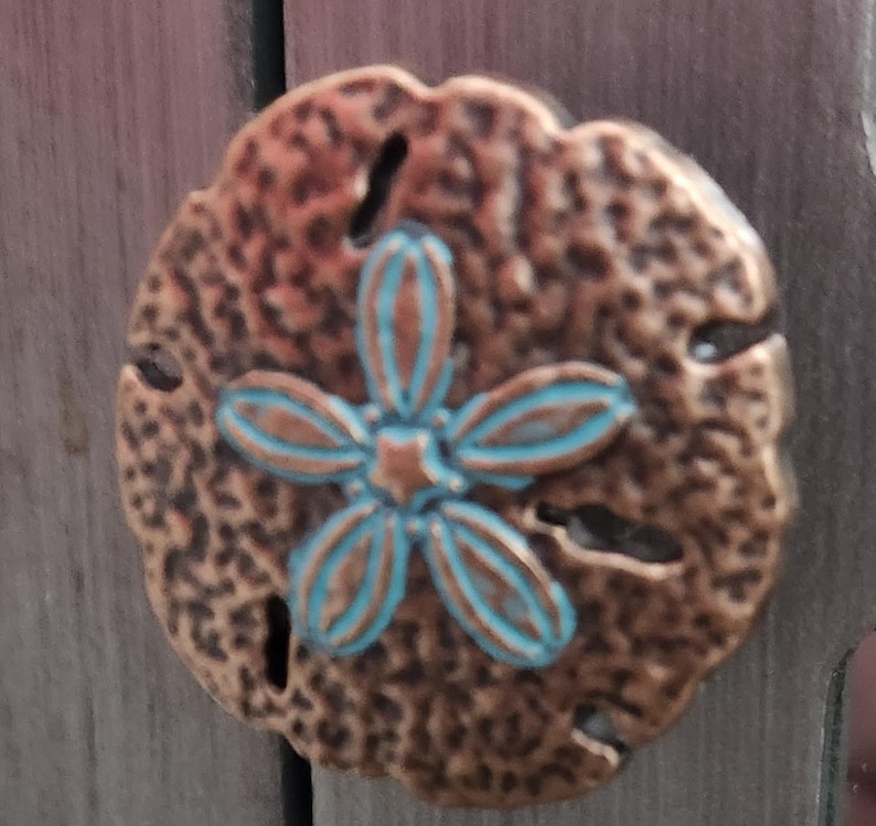 May include: A bronze-colored sand dollar-shaped decorative element with a turquoise and bronze flower design in the center. The sand dollar has a textured surface and several small holes. The design is likely for home decor.