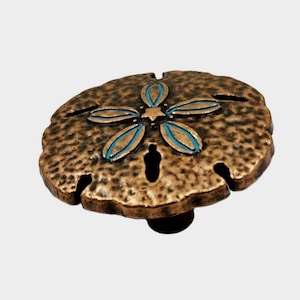 May include: Bronze-colored sand dollar cabinet knob with a textured surface. The knob features a turquoise and bronze flower design in the center and is mounted on a dark brown base. This decorative hardware adds a coastal touch to any furniture.