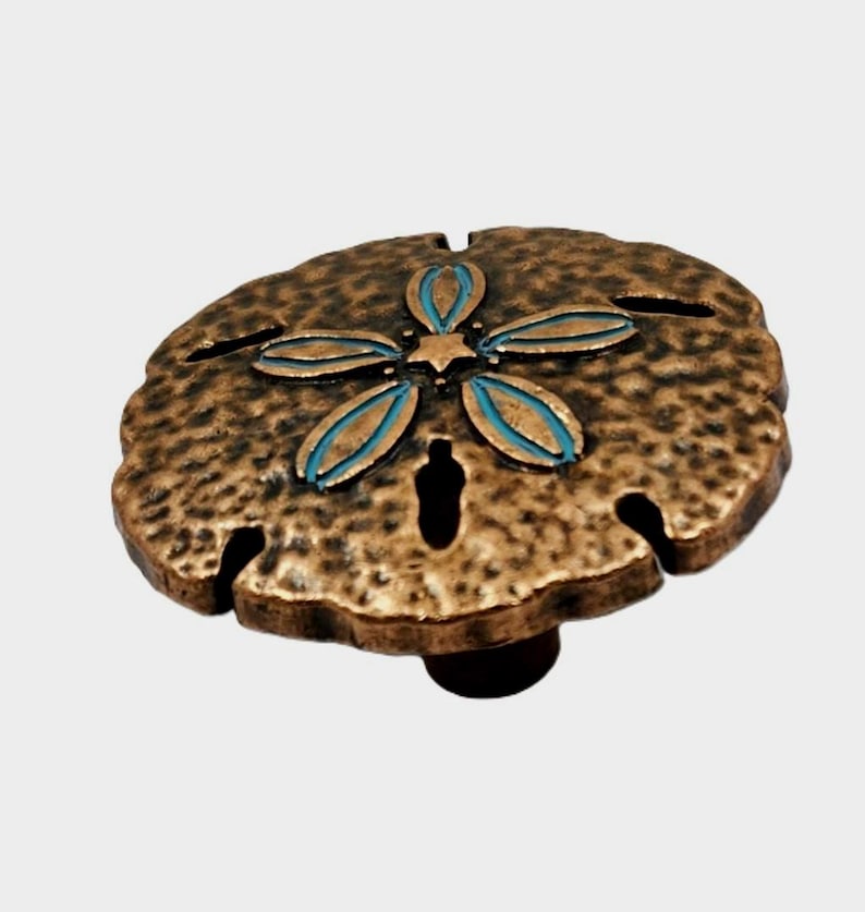 May include: Bronze-colored sand dollar cabinet knob with a textured surface. The knob features a turquoise and bronze flower design in the center and is mounted on a dark brown base. This decorative hardware adds a coastal touch to any furniture.