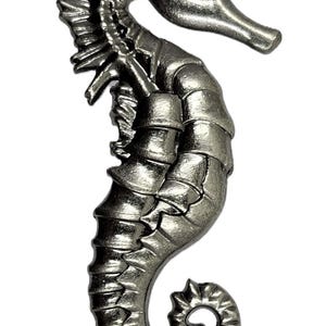May include: A silver-colored seahorse figurine with intricate details. The seahorse is curled in a traditional pose with its tail wrapped around its body.
