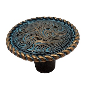May include: An oval cabinet knob with a distressed turquoise and bronze finish. The knob features a detailed floral and scroll design within a rope-like border, mounted on a dark brown base. A decorative accent for furniture.
