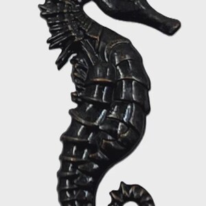 May include: A dark bronze-toned seahorse figurine featuring a segmented body, a prominent dorsal fin along the back, and a curled tail. The surface displays a metallic finish with visible ridges and textures that define the creature's anatomy.