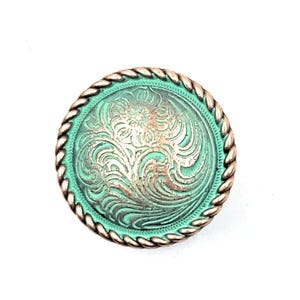 May include: A round, green metal button with a rope-like border. The button has a silver-colored, engraved floral design.
