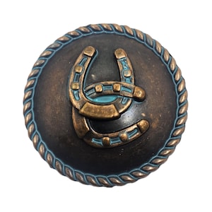 May include: A round button with a dark bronze finish featuring a raised horseshoe design in the center. The horseshoe is accented with light blue enamel details. The edge of the button is decorated with a textured rope pattern in bronze.