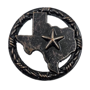May include: A circular metal emblem featuring a dark bronze finish with a distressed texture. The design depicts the outline of the state of Texas enclosed within a border of twisted barbed wire, with a prominent five-pointed star in the center.