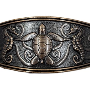 May include: An oval-shaped, bronze-toned cabinet pull featuring a sea life design. The central element is a turtle, flanked by seahorses and seashells. The design is raised and detailed, with a dark background and a lighter bronze finish on the raised elements.