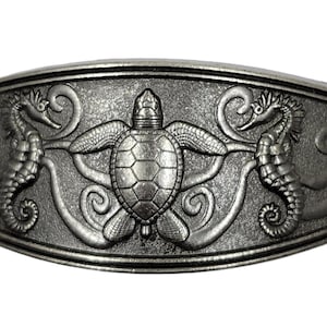 May include: A silver-toned metal belt buckle with a detailed design of a sea turtle surrounded by two seahorses. The sea turtle has a textured shell and the seahorses have intricate details.