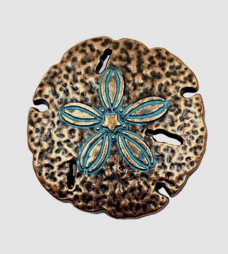 May include: Bronze-colored sand dollar with a turquoise and gold floral design in the center. The sand dollar has a textured surface and several small holes around the edges. A decorative coastal accent.
