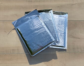 May include: Three silver bubble mailers, also known as padded envelopes, are stacked on top of each other on a light wood surface.
