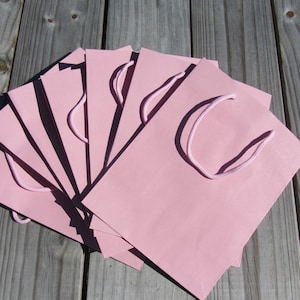 May include: A stack of pink paper gift bags with white rope handles. The bags are arranged in a fan shape on a wooden surface.