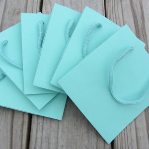 May include: A stack of six light blue gift bags with white rope handles. The bags are folded and stacked on a wooden surface.