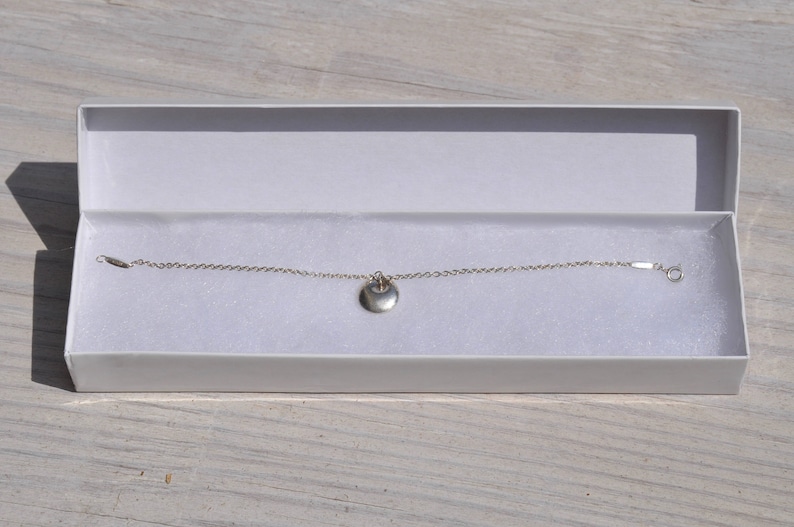 May include: A silver chain bracelet with a small, round pendant. The bracelet is in a white box with a white background.