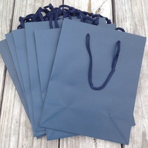 May include: A stack of dark blue paper gift bags with black handles. The bags are all the same size and are arranged in a neat pile.