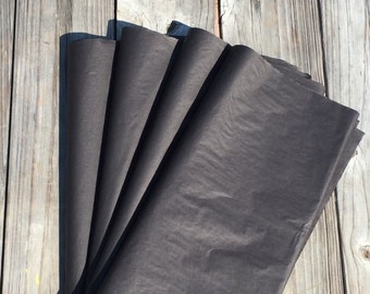 May include: A stack of six sheets of black tissue paper. The paper is folded in a fan shape and is lying on a wooden surface.