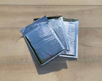 May include: Three silver bubble mailers, also known as padded envelopes, are stacked on top of each other on a light brown wooden surface.