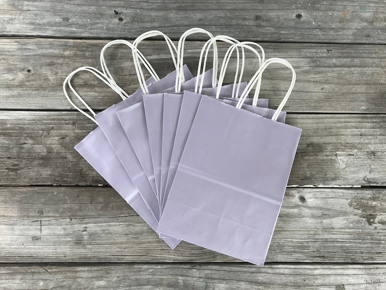 May include: A set of eight lavender-colored paper gift bags with white handles. The bags are arranged in a fan shape on a wooden surface.