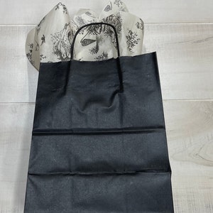 Może przedstawiać: Czarna papierowa torba prezentowa z białą podszewką z papieru tiulowgo i czarnym uchwytem. Papier tiulowy ma wz&oacute;r kwiatowy.