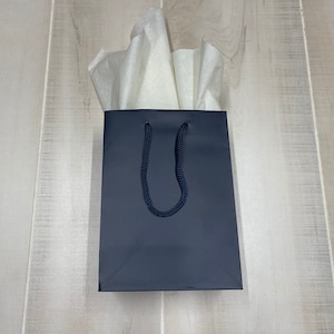 May include: A dark blue gift bag with a black rope handle. The bag is empty and has white tissue paper inside.