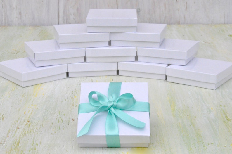 May include: A white gift box with a light blue ribbon tied in a bow. The box is sitting on a white wooden surface.