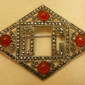 May include: A silver brooch with a diamond shape design. The brooch features the letters "D" and "C" in the center, surrounded by a border of small stones. The brooch is accented with four red carnelian stones.