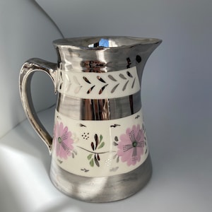 May include: Silver-plated pitcher with a handle and spout. The pitcher features a cream-colored band with pink floral designs and a silver band at the top and bottom. The handle and spout are also silver-plated.
