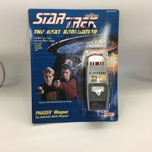 May include: A silver Star Trek: The Next Generation Phaser toy with a red and black button panel. The toy is packaged in a blue and white cardboard box with images of the characters from the show. The box also includes the text "PHASER™ Weapon" and "The Authentic Battle Weapon!"