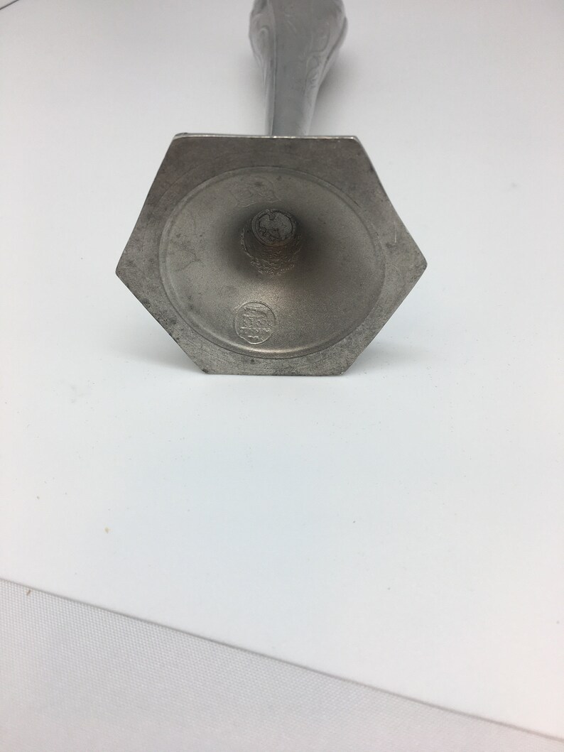 Pu&ograve; includere: Un candeliere color peltro con base esagonale e stelo sottile e decorato. La base presenta un'indentazione circolare con un piccolo stemma. Il candeliere &egrave; in metallo e ha un aspetto vintage.