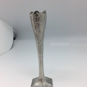 Pu&ograve; includere: Un vaso a bocciolo color argento con un motivo floreale. Il vaso ha una base quadrata svasata e un corpo sottile e allungato. La parte superiore del vaso ha un bordo smerlato. Il motivo floreale &egrave; inciso nel metallo.