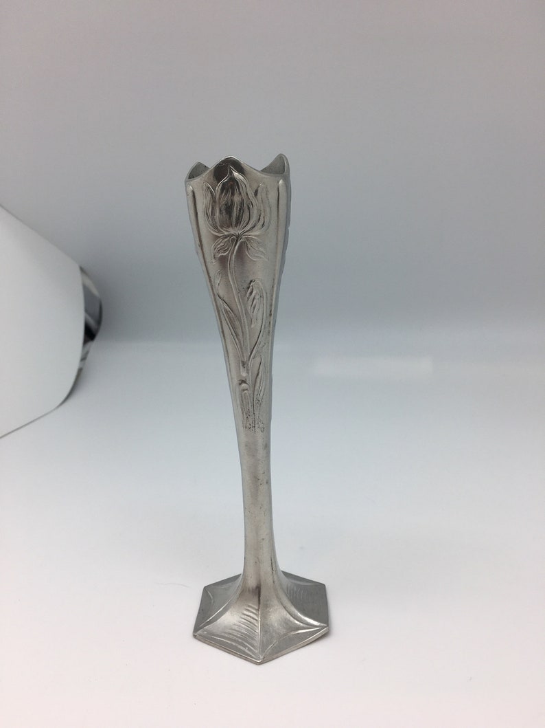 Pu&ograve; includere: Un vaso a bocciolo color argento con un motivo floreale. Il vaso ha una base quadrata svasata e un corpo sottile e allungato. La parte superiore del vaso ha un bordo smerlato. Il motivo floreale &egrave; inciso nel metallo.