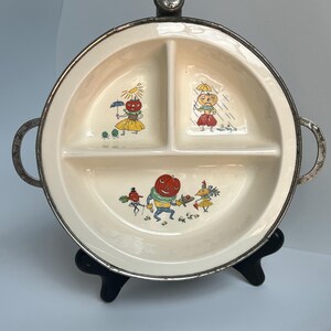 May include: Vintage divided serving dish with a cream-colored interior and a silver-toned metal rim and handles. The dish features three sections, each decorated with whimsical illustrations of anthropomorphic vegetables. The dish is displayed on a black stand.