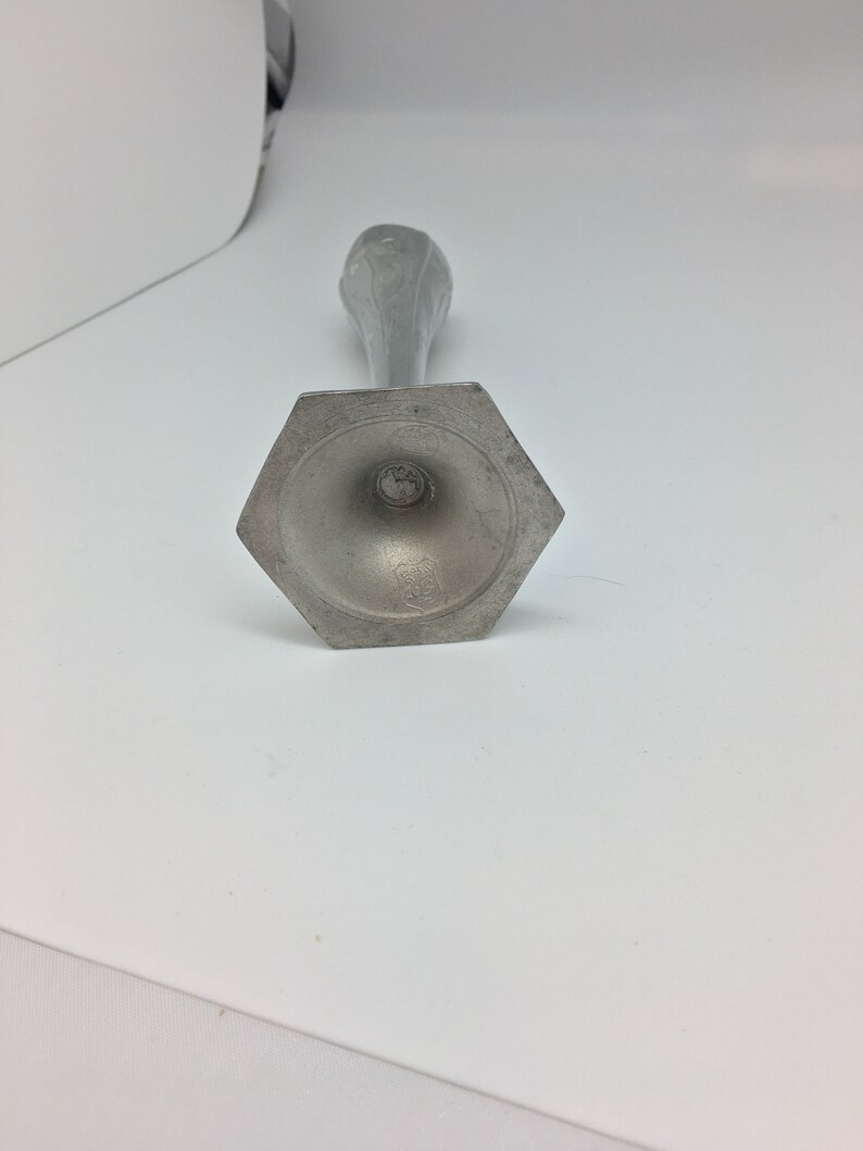 Pu&ograve; includere: Un portacandele color argento con base esagonale e stelo sottile. La parte superiore del supporto presenta un motivo floreale. Il portacandele &egrave; in metallo e si trova su una superficie bianca.