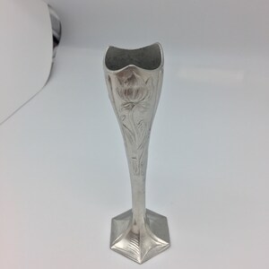 Pu&ograve; includere: Un vaso portafiori argentato con base esagonale e corpo affusolato. Il vaso presenta un motivo floreale in rilievo e una parte superiore svasata e smerlata. L'altezza &egrave; di circa 18 cm.