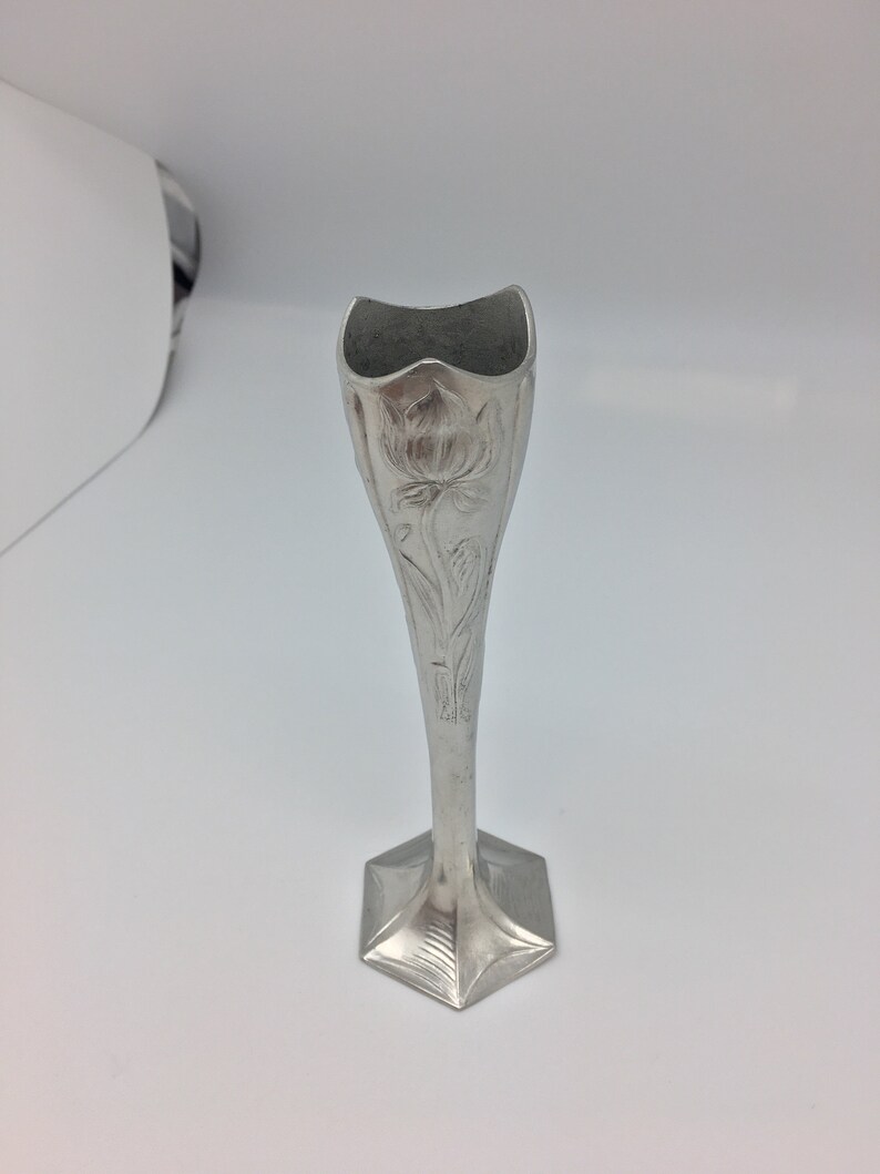 Pu&ograve; includere: Un vaso portafiori argentato con base esagonale e corpo affusolato. Il vaso presenta un motivo floreale in rilievo e una parte superiore svasata e smerlata. L'altezza &egrave; di circa 18 cm.