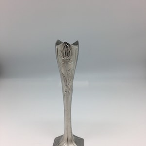 Pu&ograve; includere: Un vaso argentato e sottile con un motivo floreale. Il vaso ha una parte superiore svasata e un corpo lungo e affusolato. La base ha una forma geometrica. Il motivo floreale &egrave; inciso nel metallo.