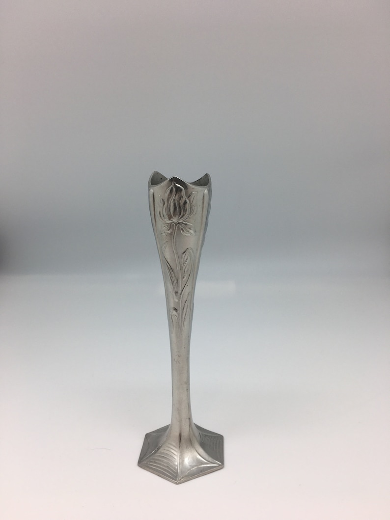 Pu&ograve; includere: Un vaso argentato e sottile con un motivo floreale. Il vaso ha una parte superiore svasata e un corpo lungo e affusolato. La base ha una forma geometrica. Il motivo floreale &egrave; inciso nel metallo.