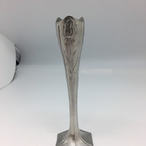 Pu&ograve; includere: Un vaso argentato e slanciato con un motivo floreale. Il vaso ha una parte superiore svasata e un corpo lungo e affusolato. La base &egrave; esagonale. Il motivo floreale &egrave; una rosa. Il vaso &egrave; probabilmente in metallo.