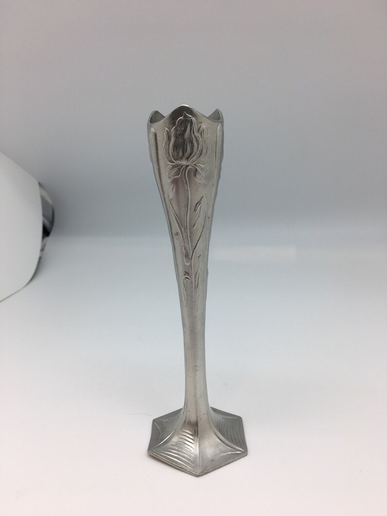 Pu&ograve; includere: Un vaso argentato e slanciato con un motivo floreale. Il vaso ha una parte superiore svasata e un corpo lungo e affusolato. La base &egrave; esagonale. Il motivo floreale &egrave; una rosa. Il vaso &egrave; probabilmente in metallo.