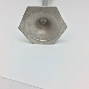 Pu&ograve; includere: Un candelabro in metallo color argento con base esagonale. La base presenta un disegno circolare attorno al centro e un piccolo stemma. Lo stelo del candelabro &egrave; sottile e si assottiglia verso una parte superiore decorativa.