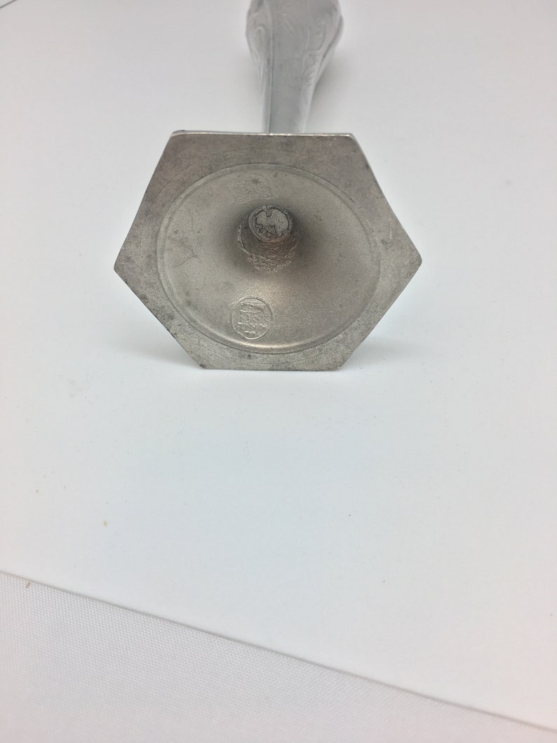 Pu&ograve; includere: Un candelabro in metallo color argento con base esagonale. La base presenta un disegno circolare attorno al centro e un piccolo stemma. Lo stelo del candelabro &egrave; sottile e si assottiglia verso una parte superiore decorativa.