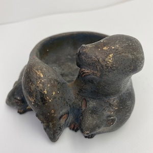May include: A black ceramic ashtray shaped like three mice huddled together. The ashtray has a gold-colored glaze and a small, round bowl in the center.
