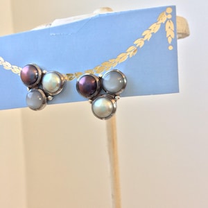 May include: A pair of silver-toned stud earrings, each featuring three iridescent, round stones in shades of purple, white, and gray. The earrings are displayed on a light blue card with a gold leaf design.
