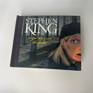 May include: A hardcover book titled "The Girl Who Loved Tom Gordon" by Stephen King. The cover features a forest scene and a portrait of a person with a surprised expression. The book is a Pop-up Book.