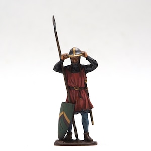 May include: A miniature figurine of a medieval knight, clad in chainmail and a red tunic, holding a spear and a shield. The knight is adjusting their helmet. The shield is green with a yellow stripe. The figurine is on a brown base.