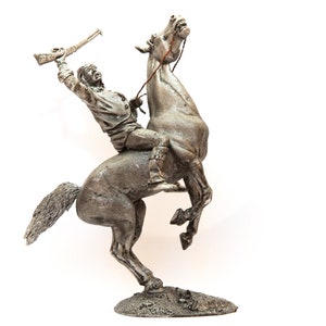 Op de afbeelding: Een zilverkleurig beeldje van een indiaanse krijger die een opspringend paard berijdt. De krijger houdt een geweer in zijn rechterhand en draagt een verenhoofdtooi.