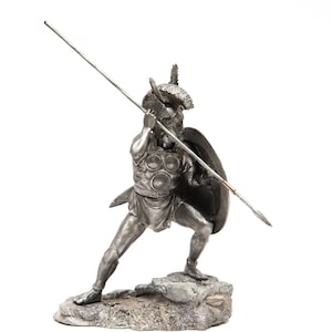 May include: A silver figurine of a warrior holding a spear and shield. The warrior is wearing a helmet and armor. The figurine is standing on a rocky base.