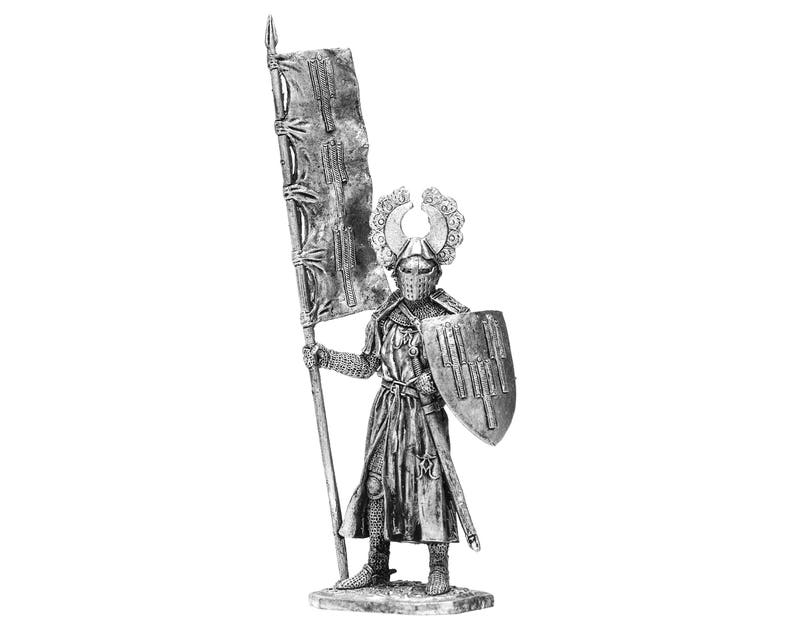 May include: A silver miniature figure of a knight in armour holding a flag and a shield. The knight is wearing a helmet with a crest and a long, flowing robe. The flag is white with a red cross. The shield is round and has a red and white design.