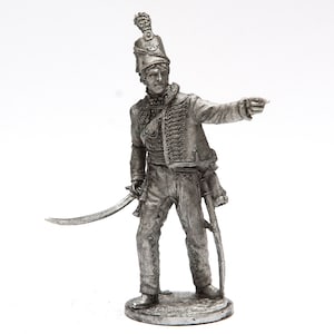 Puede incluir: Una figurita de metal pintada de plata de un soldado que lleva un sombrero alto con una pluma, una chaqueta con cuello de piel y sostiene una espada y un rifle. El soldado está de pie sobre una base redonda.
