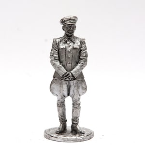 May include: A silver-colored metal figurine of a man in a military uniform. He is wearing a cap with a visor and a jacket with buttons. He is standing with his hands clasped in front of him.