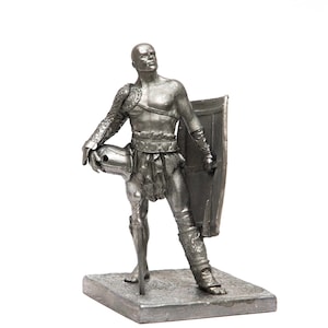 Puede incluir: Una figurita de plata de un gladiador romano sosteniendo un escudo y una espada. El gladiador lleva armadura y un taparrabos. La figurita está de pie sobre una base cuadrada.